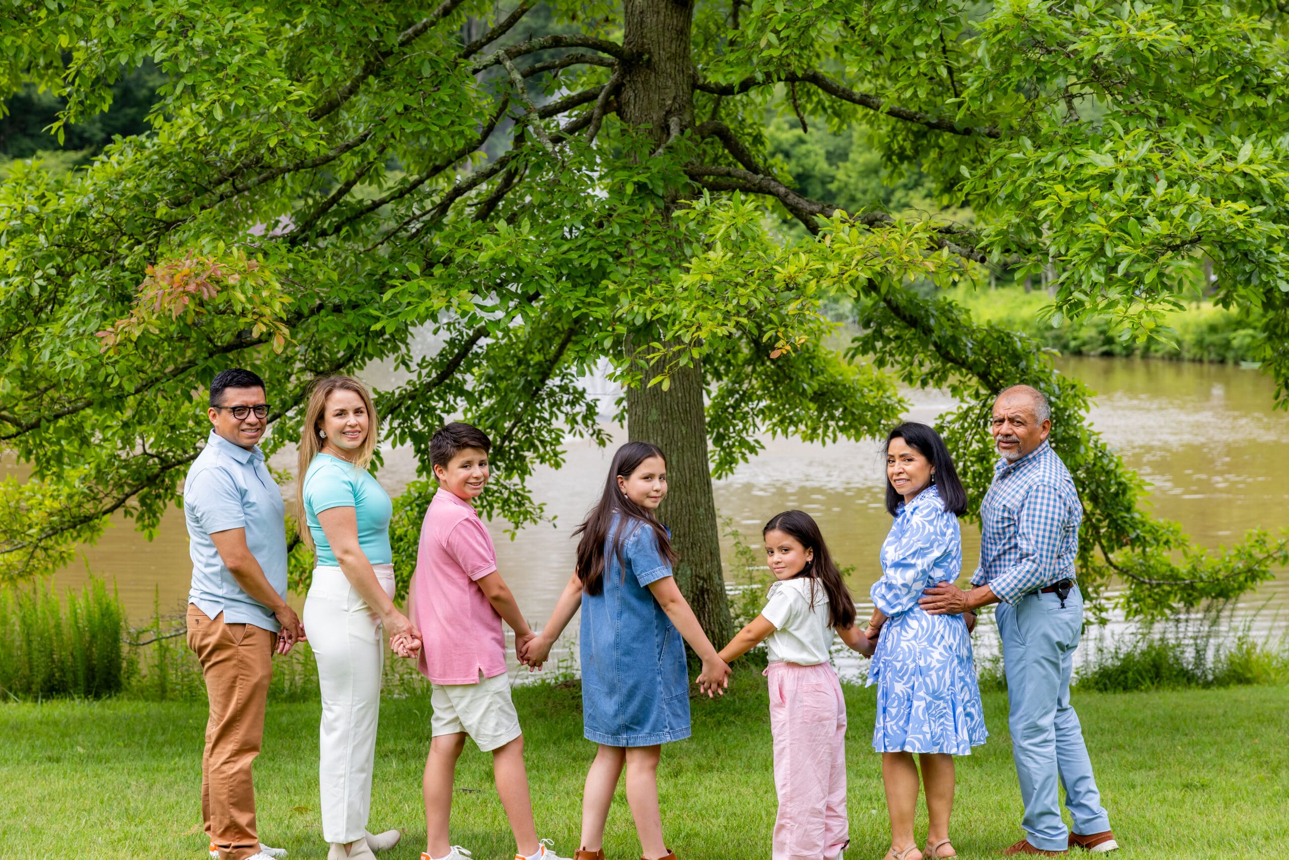 Three generations holding hands in a line under a large oak tree by the pond at Meadowlark Botanical Gardens – captured by Capture Joy Photography, Northern Virginia family photographer.