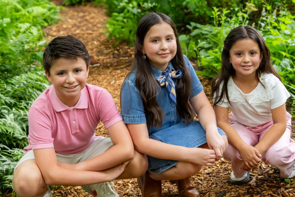 Two sisters and their brother sitting in the garden surrounded by trees and mulch, smiling naturally – captured by Capture Joy Photography, Northern Virginia family photographer.