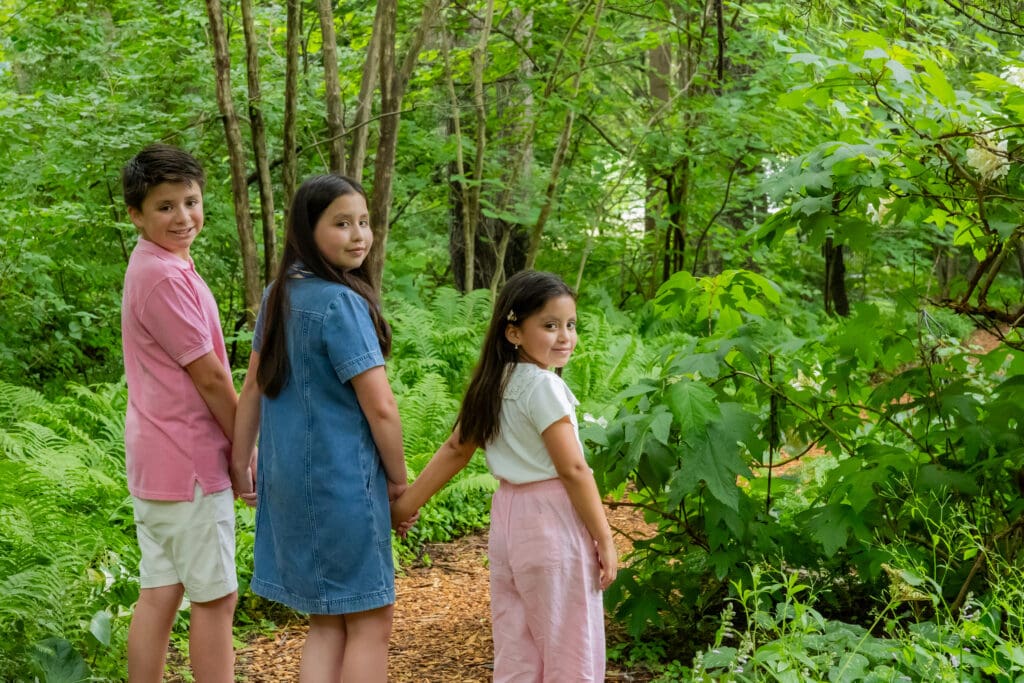 Three siblings standing side by side in the woods at Meadowlark Botanical Gardens, showing candid expressions – captured by Capture Joy Photography, Northern Virginia family photographer.