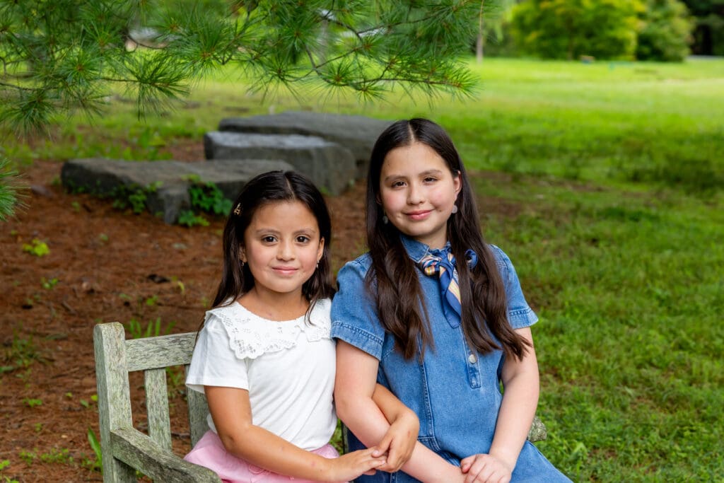 Two sisters seated together on a bench, smiling sweetly in the gardens – captured by Capture Joy Photography, Northern Virginia family photographer.