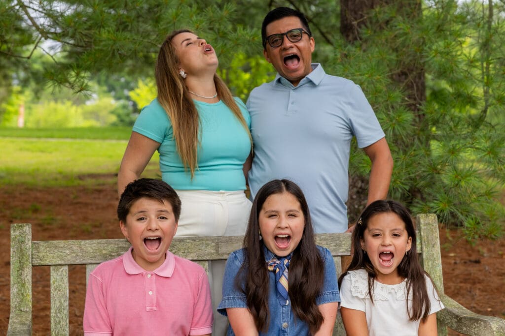 Parents and three children sitting on a wooden bench at Meadowlark Botanical Gardens making silly faces and laughing together – captured by Capture Joy Photography, Northern Virginia family photographer.