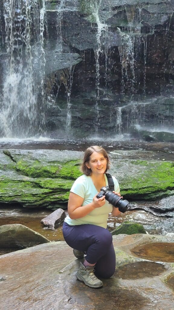 Photographer kneeling on rocks near a waterfall holding Canon R8 with Lemur Strap resting across shoulder, enjoying outdoor adventure photography – captured by Capture Joy Photography, Northern Virginia family photographer.