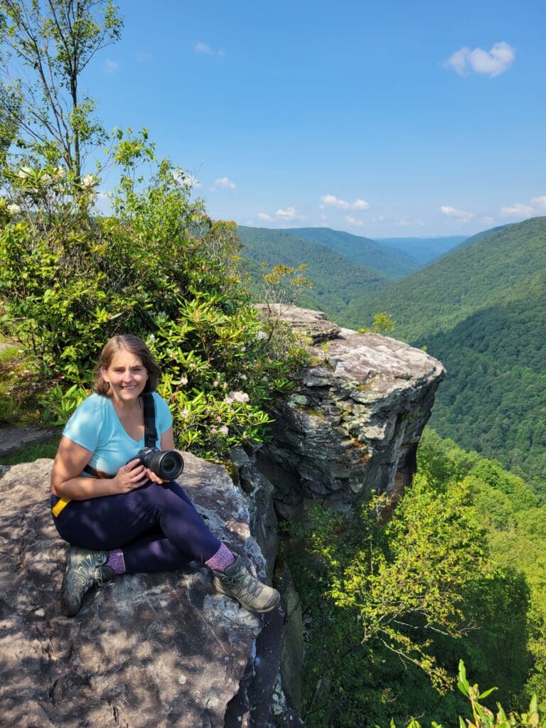 Photographer smiling and holding Canon R8 with Lemur Strap while sitting on a rocky ledge overlooking lush green mountains – captured by Capture Joy Photography, Northern Virginia family photographer.