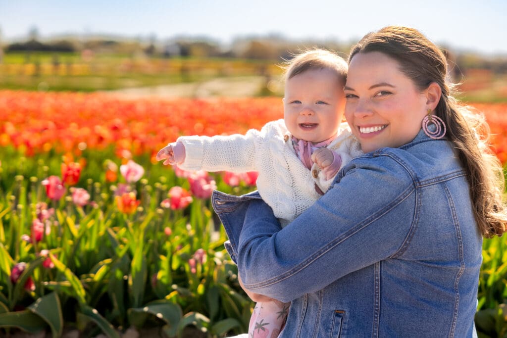 Mother holding baby girl close, both smiling with a sea of red and pink tulips behind them – captured by Capture Joy Photography, Northern Virginia family photographer.