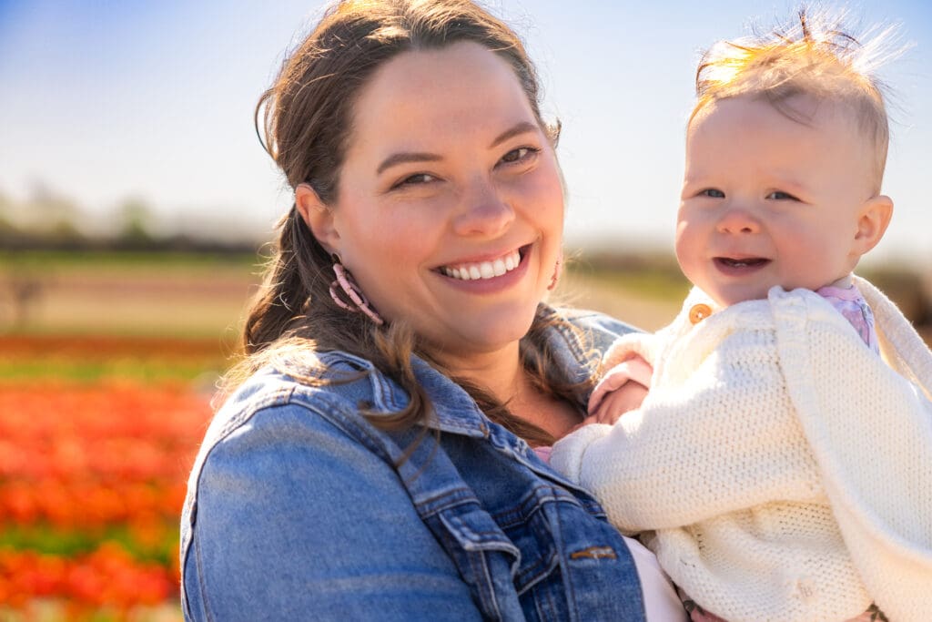 Happy mother holding baby girl on her lap, both smiling with tulips in the distance – captured by Capture Joy Photography, Northern Virginia family photographer.